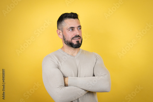 Handsome man with beard wearing sweater over yellow background looking to the side with arms crossed convinced and confident