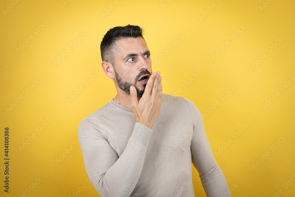 Handsome man with beard wearing sweater over yellow background covering mouth with hand, shocked and afraid for mistake. surprised expression Handsome man with beard wearing sweater over yellow background covering mouth with hand, shocked and afraid for mistake. surprised expression