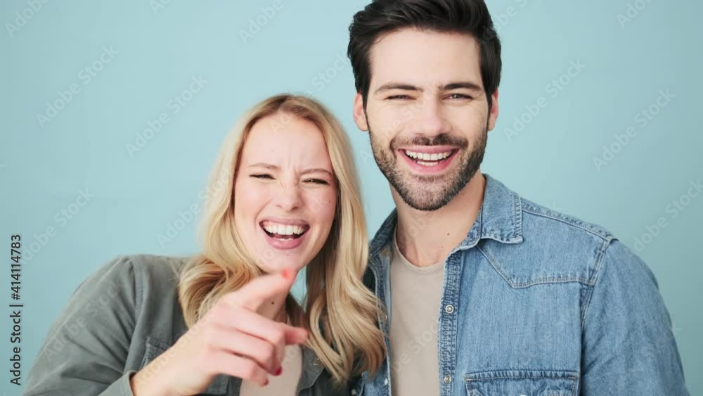 Pleased couple man and woman are pointing to the camera and laughing standing isolated over blue background in studio Pleased couple man and woman are pointing to the camera and laughing standing isolated over blue background in studio
