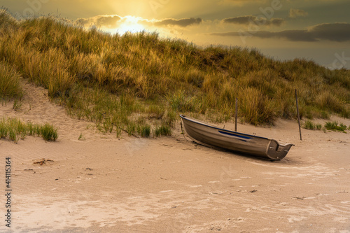 Fishing boat lies on the beach in the sunset in front of a dune