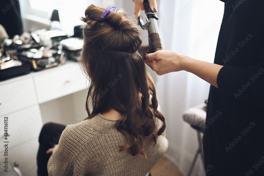 Hair stylist prepares beautiful young woman for event, makes curls hairstyle with a curling iron for client in beauty salon. Long beautiful light brown natural hair. Hair stylist prepares beautiful young woman for event, makes curls hairstyle with a curling iron for client in beauty salon. Long beautiful light brown natural hair.
