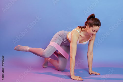 European lady on all fours with resistance band on knees, take one leg to side, woman works out indoor isolated over color background, looking down, wearing stylish sportswear. © sementsova321