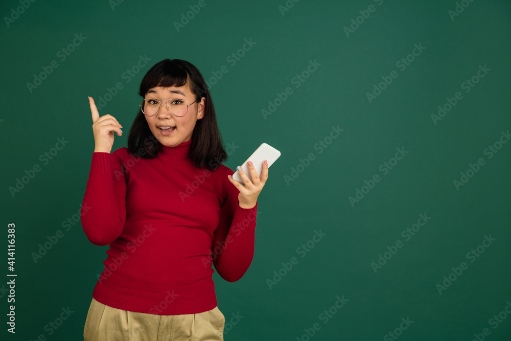 Pointing up with phone. East asian young beautiful woman's portrait on green studio background with copyspace. Brunette female model. Concept of human emotions, facial expression, sales, ad, fashion.