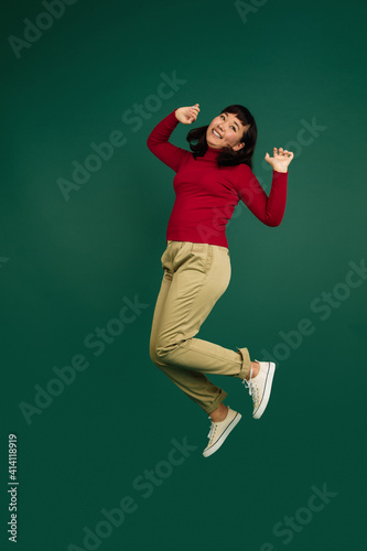 Jumping high cheerful. East asian young beautiful woman's portrait on green studio background with copyspace. Brunette female model. Concept of human emotions, facial expression, sales, ad, fashion.