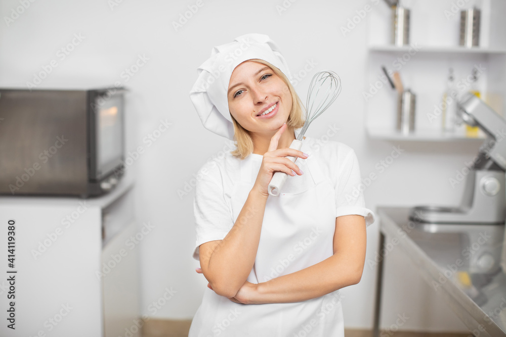 Close up portrait of young beautiful smiling blond girl, professional pastry chef, wearing white uniform, apron and chef's hat, posing to camera with cook whisk, standing in the kitchen