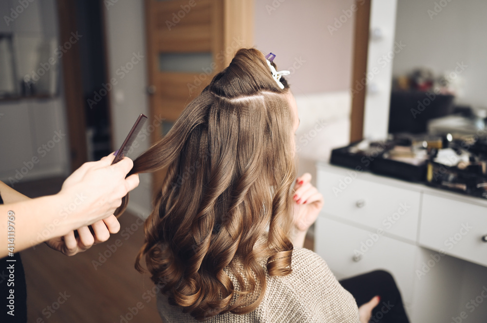 Fototapeta premium Hair stylist prepares beautiful young woman for event, makes curls hairstyle with a curling iron for client in beauty salon. Long beautiful light brown natural hair.