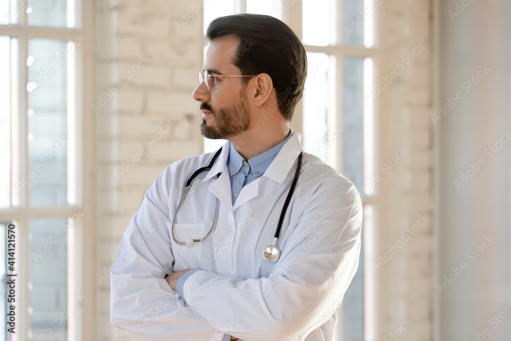 Thoughtful young male doctor in eyewear and medical coat standing in clinic office with folded arms, looking outside in distance, thinking of career opportunities or professional challenges. Thoughtful young male doctor in eyewear and medical coat standing in clinic office with folded arms, looking outside in distance, thinking of career opportunities or professional challenges.