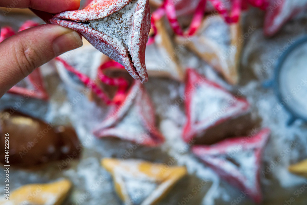 Hand showing pink colorful hamantash cookie with more hamantaschen in background. Jewish traditional pastry for Purim (Jewish carnival holiday). Homemade gluten free cookie filled with chocolate.
