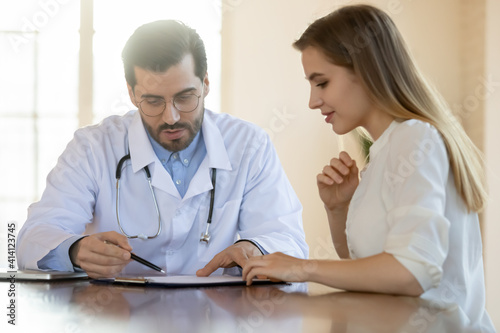 Serious male family doctor explaining medical insurance agreement to interested young female patient at meeting. Focused woman discussing health test results with caring gp in clinic office.