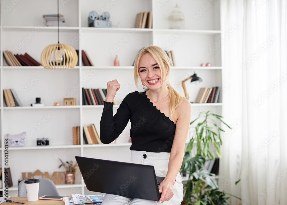 Young blond woman, wearing black top and white pants, sitting on office table, holding laptop, being happy, smiling, Office manager at her workplace. Female working on computer in company. Young blond woman, wearing black top and white pants, sitting on office table, holding laptop, being happy, smiling, Office manager at her workplace. Female working on computer in company.