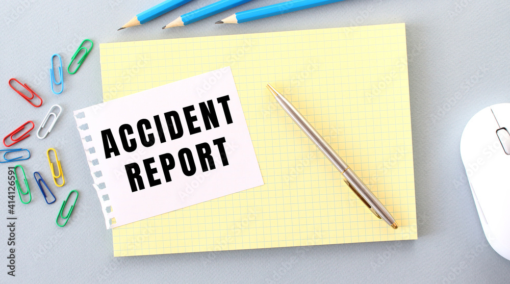 ACCIDENT REPORT is written on a piece of paper that lies on a notebook next to office supplies. ACCIDENT REPORT is written on a piece of paper that lies on a notebook next to office supplies.