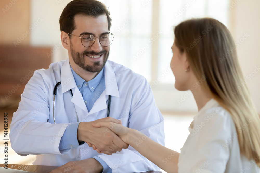 Smiling young handsome male gp doctor in eyeglasses shaking hands with happy female patient, welcoming at visit, making agreement or signing deal at consultation, getting acquainted in clinic office. Smiling young handsome male gp doctor in eyeglasses shaking hands with happy female patient, welcoming at visit, making agreement or signing deal at consultation, getting acquainted in clinic office.