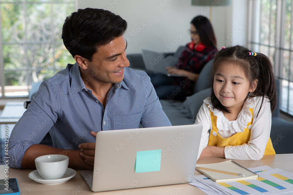 Mixed race family staying together, Caucasian father sitting and working at desk and teaching half-race little cute daughter white Asian mother using laptop computer on sofa. Idea for work at home Mixed race family staying together, Caucasian father sitting and working at desk and teaching half-race little cute daughter white Asian mother using laptop computer on sofa. Idea for work at home