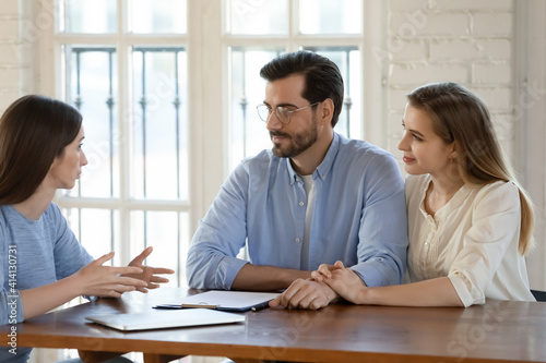 Happy young family couple visiting female real estate agent, discussing agreement details. Professional financial advisor discussing savings investment with married clients at office meeting.