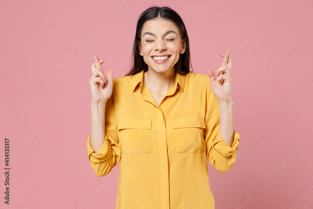 Young brunette cute latin woman 20s wearing yellow casual shirt waiting for special moment, keeping fingers crossed, making wish with closed eyes isolated on pastel pink background studio portrait. Young brunette cute latin woman 20s wearing yellow casual shirt waiting for special moment, keeping fingers crossed, making wish with closed eyes isolated on pastel pink background studio portrait.