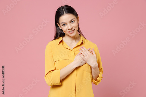 Young positive thankful soft-hearted kind good brunette nice attractive latin woman 20s wearing yellow casual shirt put folded hands on heart isolated on pastel pink color background studio portrait.