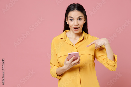 Young brunette attractive latin shocked surprised wondered confused woman 20s in yellow shirt point index finger on mobile cell phone chatting isolated on pastel pink color background studio portrait.