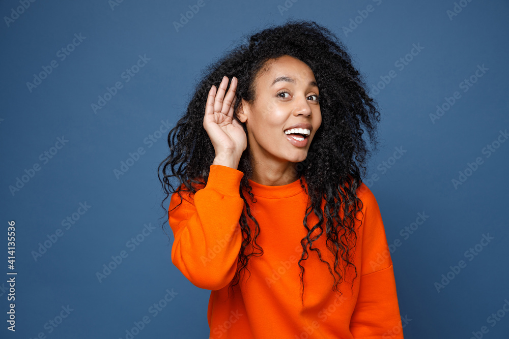 Excited curious young african american woman in casual basic bright orange sweatshirt try to hear you overhear listening intently looking camera isolated on blue color background studio portrait. Excited curious young african american woman in casual basic bright orange sweatshirt try to hear you overhear listening intently looking camera isolated on blue color background studio portrait.