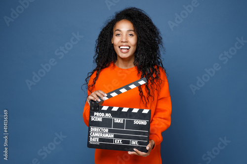 Photos Laughing young african american woman in casual basic bright orange sweatshirt standing holding classic black film making clapperboard looking camera isolated on blue color background studio portrait