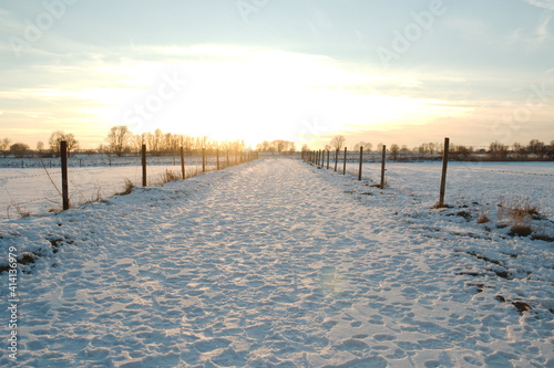 Dutch landscape in winter with snow  a country road and a meadow with pasture posts and barbed wire. Trees on the horizon. Sunset in province Gelderland. With copy space.