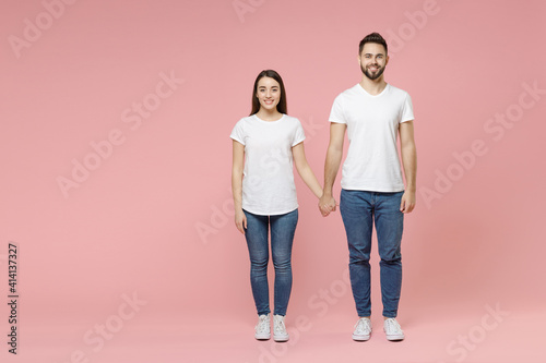 Full body length young cheerful couple two friends bearded man brunette woman in white basic blank print design t-shirts jeans standing posing isolated on pastel pink color background studio portrait.