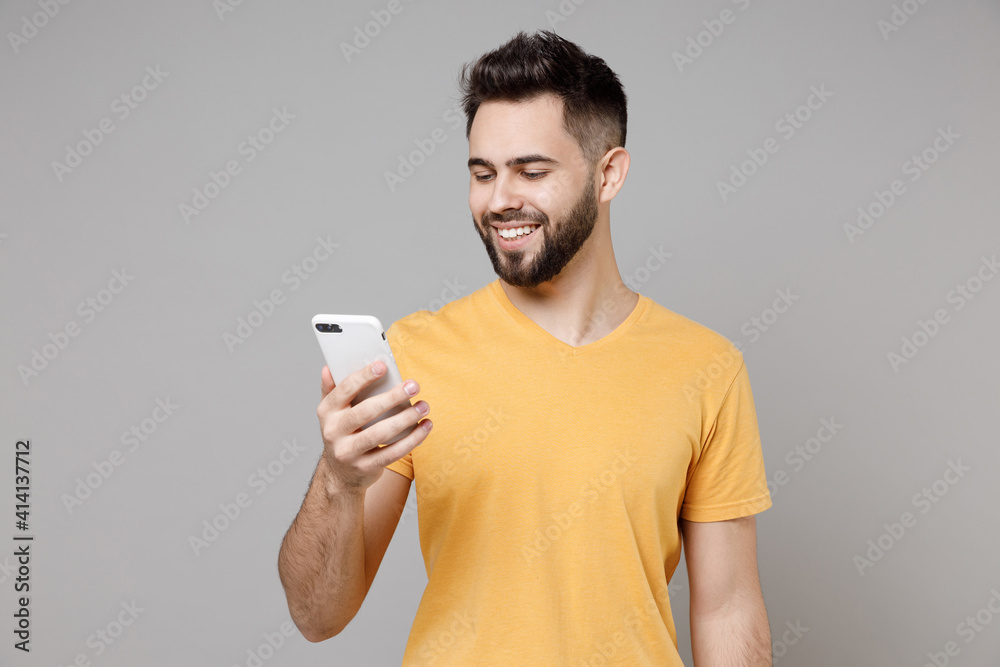Young caucasian smiling bearded attractive man 20s wear casual yellow basic t-shirt hold mobile cell phone chat in social network isolated on grey background studio portrait People lifestyle concept. Young caucasian smiling bearded attractive man 20s wear casual yellow basic t-shirt hold mobile cell phone chat in social network isolated on grey background studio portrait People lifestyle concept.