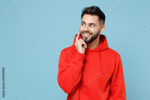 Young caucasian smiling pensive wistful dreamful bearded man 20s in casual red orange hoodie look aside prop up face with finger isolated on blue background studio portrait People lifestyle concept