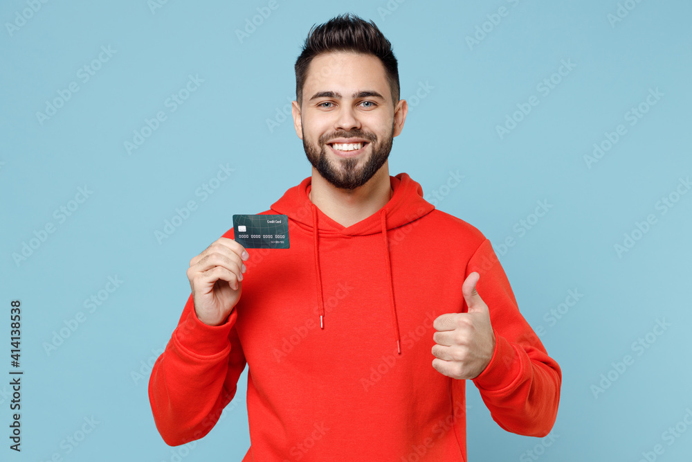 Young caucasian smiling happy satisfied rich bearded student man 20s in casual red orange hoodie holding credit bank card show thumb up gesture look camera isolated on blue background studio portrait. Young caucasian smiling happy satisfied rich bearded student man 20s in casual red orange hoodie holding credit bank card show thumb up gesture look camera isolated on blue background studio portrait.
