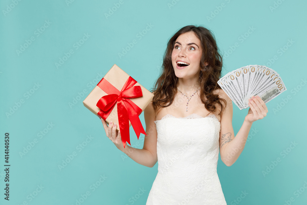 Εκτύπωση καμβά Shocked bride young woman in white wedding dress hold present box with gift ribbon bow fan of cash money in dollar banknotes isolated on blue turquoise background