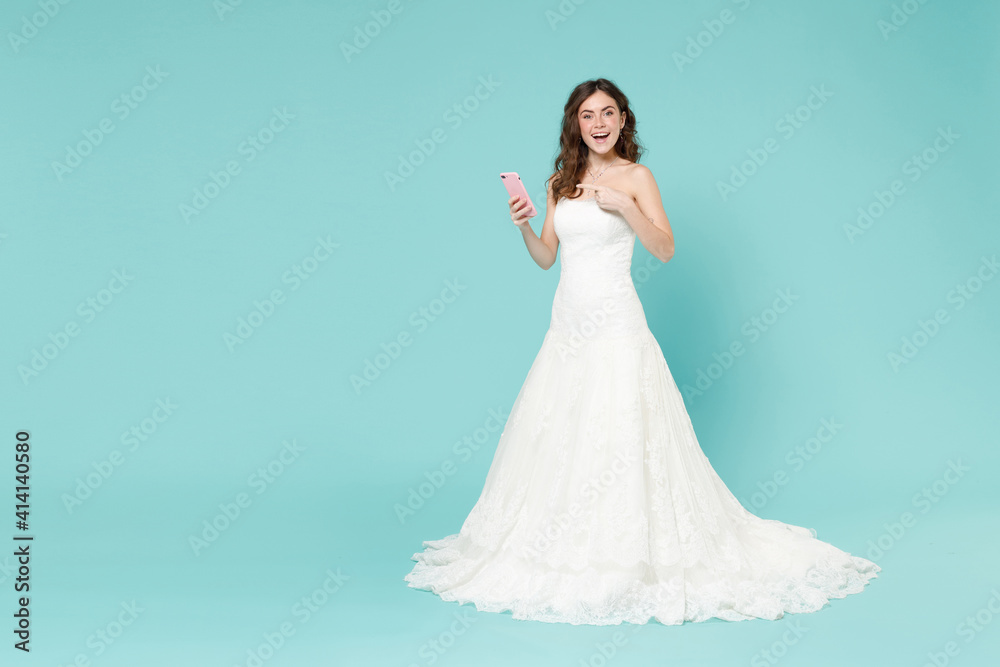 Full length excited bride young woman 20s in white wedding dress pointing index finger on mobile cell phone isolated on blue turquoise background studio portrait. Ceremony celebration party concept. Full length excited bride young woman 20s in white wedding dress pointing index finger on mobile cell phone isolated on blue turquoise background studio portrait. Ceremony celebration party concept.
