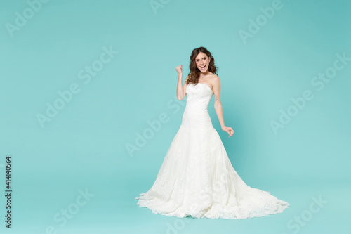 Full length excited happy bride young woman in white wedding dress doing winner gesture celebrating clenching fists say yes isolated on blue turquoise background. Ceremony celebration party concept.