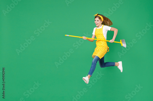 Full length side view of funny little kid girl housewife 12-13 in yellow apron jumping running hold broom doing housework isolated on green background children studio portrait. Housekeeping concept.