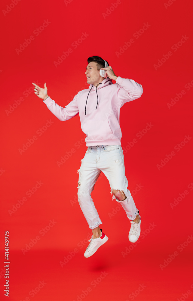 Jumping high with headphones. Caucasian man's portrait isolated on red background with copyspace. Handsome male model in street style. Concept of human emotions, facial expression, sales, ad, fashion.