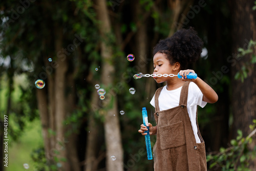 Little African American curly hair girl in casual clothing holding bubble wand blowing bubbles playing alone at outdoor. photo