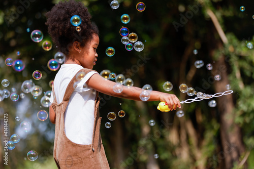 Valokuvatapetti Little African American curly hair girl in casual clothing holding bubble wand blowing bubbles playing alone at outdoor
