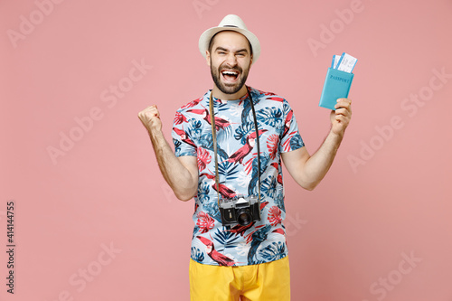 Happy young traveler tourist man in summer clothes hat hold passport ticket boarding pass doing winner gesture isolated on pink background. Passenger traveling on weekends. Air flight journey concept.