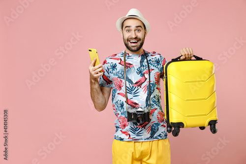 Excited young traveler tourist man in summer clothes hat hold suitcase using mobile phone booking taxi hotel isolated on pink background. Passenger traveling on weekends. Air flight journey concept.