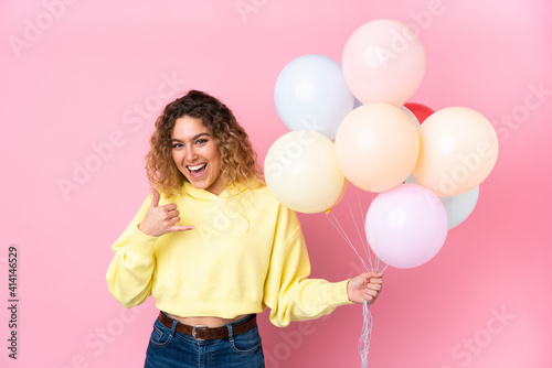 Young blonde woman with curly hair catching many balloons isolated on pink background making phone gesture