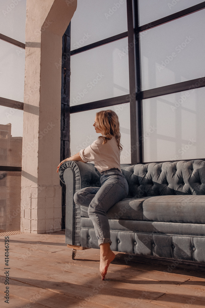 Obraz premium young woman in white shirt and jeans is sitting and relaxing with smile in sunlights in blue sofa near panoramic windows background, lifestyle concept , free space