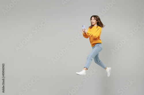 Obraz na płótnie Full length of young fun smiling caucasian student woman 20s wearing knitted yellow sweater jumping high hold mobile cell phone chatting typing sms isolated on grey color background studio portrait