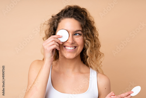 Young blonde woman with curly hair isolated on beige background with cotton pad for removing makeup from her face and smiling