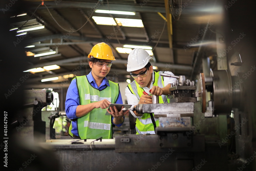 Asian engineering manager and mechanic worker in safety hard hat and reflective cloth using lathe machine inside the factory
