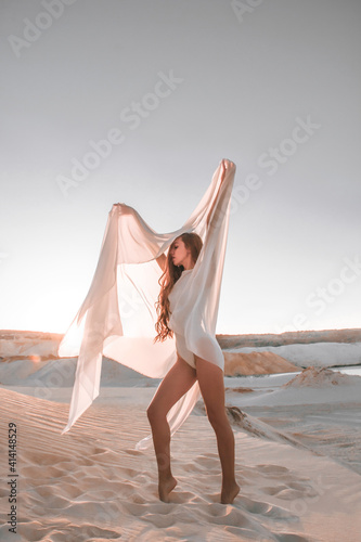 young woman is standing in gymnastic pose on the sand career with white transparent cloth in hands at sunset sky  background, lifestyle concept, free space
