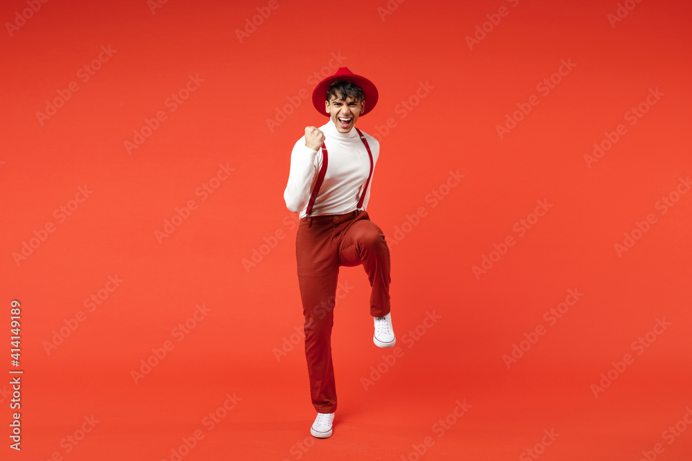 Full length of young spanish latinos successful lucky man 20s in hat white shirt trousers, suspenders do winner gesture clench fists with raised up leg isolated on red color background studio portrait Full length of young spanish latinos successful lucky man 20s in hat white shirt trousers, suspenders do winner gesture clench fists with raised up leg isolated on red color background studio portrait