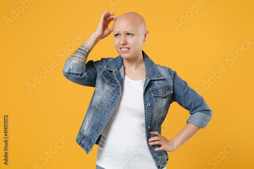 Young bald caucasian pensive thoughful troubled woman 20s without hair wearing casual denim jacket white t-shirt looking aside sctratch temple head isolated on yellow color background studio portrait. photo
