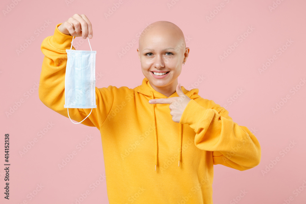 Young bald woman 20s without hair in yellow casual shirt hold point index finger on sterile face mask safe coronavirus covid-19, pandemic quarantine isolated on pastel pink background studio portrait Young bald woman 20s without hair in yellow casual shirt hold point index finger on sterile face mask safe coronavirus covid-19, pandemic quarantine isolated on pastel pink background studio portrait