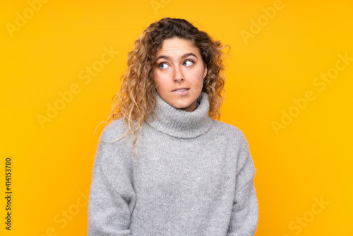 Young blonde woman with curly hair wearing a turtleneck sweater isolated on yellow background with confuse face expression © luismolinero