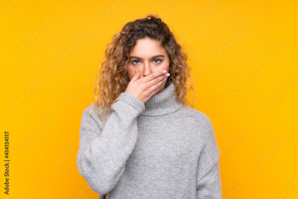 Young blonde woman with curly hair wearing a turtleneck sweater isolated on yellow background covering mouth with hands Young blonde woman with curly hair wearing a turtleneck sweater isolated on yellow background covering mouth with hands
