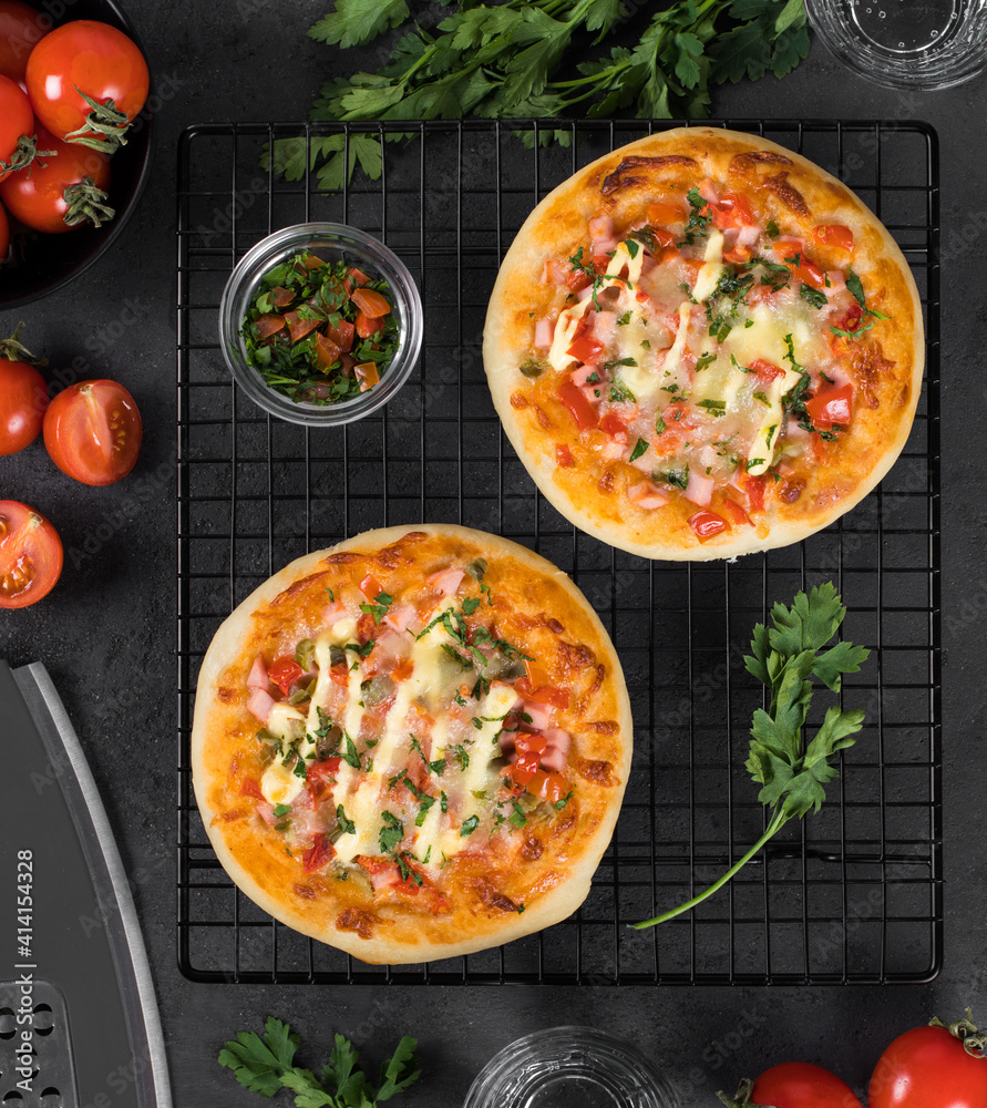 Two pies pizzas with sausage, pickles, tomatoes, mozzarella, greens on a metal stand on a dark gray background top view 