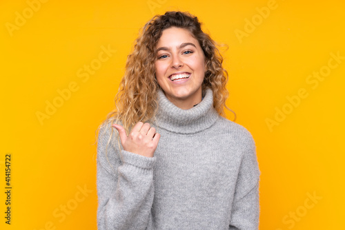 Young blonde woman with curly hair wearing a turtleneck sweater isolated on yellow background pointing to the side to present a product © luismolinero